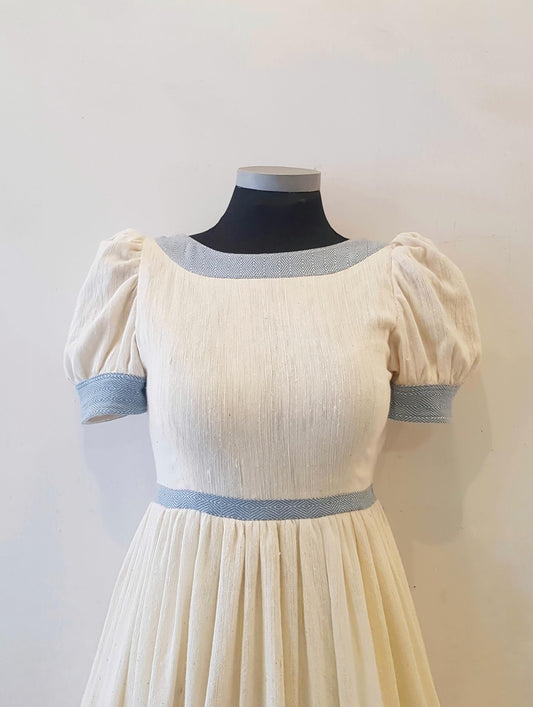 A cream-colored dress with light blue Ethiopian Outfit  on the neckline, sleeve cuffs, and waistband, displayed on a black dress form mannequin against a plain off-white wall.