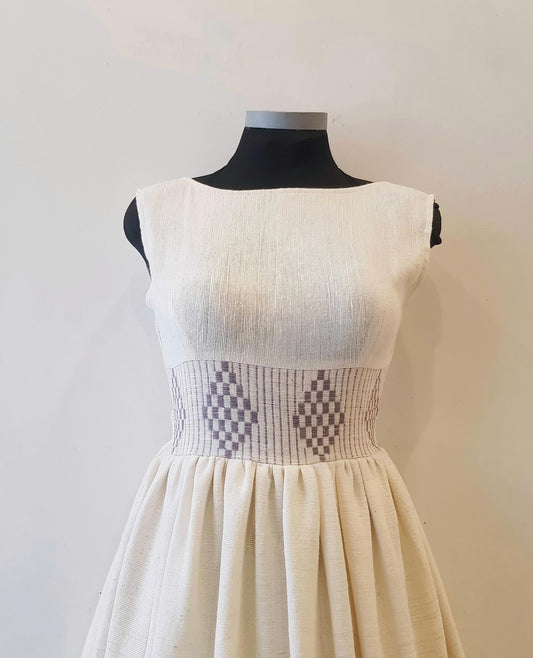 An off-white sleeveless Ethiopian Outfit displayed on a black mannequin torso against a plain light background. The dress has a textured linen-like bodice and skirt, with a smocked patterned waistband featuring gray and white diamond and checkerboard motifs.