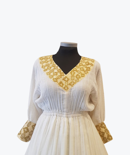 A white long-sleeve Ethiopian Outfit  displayed on a black mannequin torso against a light gray background. The dress features a V-neckline with ornate gold embroidery, gathered cuffs with matching gold embroidery, and a pleated bodice.