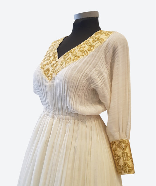 An off-white or cream pleated long-sleeve Ethiopian Outfit  displayed on a black mannequin torso. The dress features ornate gold embroidered detailing along the V-neckline and on the cuffs.