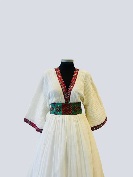 A white or cream-colored traditional Ethiopian Outfit displayed on a black mannequin torso against a light gray background. The dress features a V-neckline with red and green patterned trim, a matching patterned waist belt, and patterned trim on the wide sleeves.