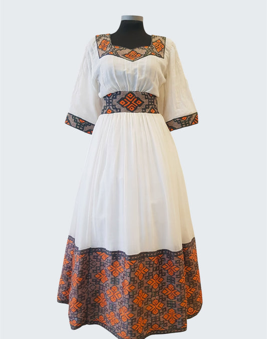 A white midi ethiopian outfit displayed on a black mannequin against a light gray background. The dress features a square neckline with orange and dark gray geometric embroidery, elbow-length sleeves with matching patterned cuffs, a patterned waistband, and a wide matching patterned hem.