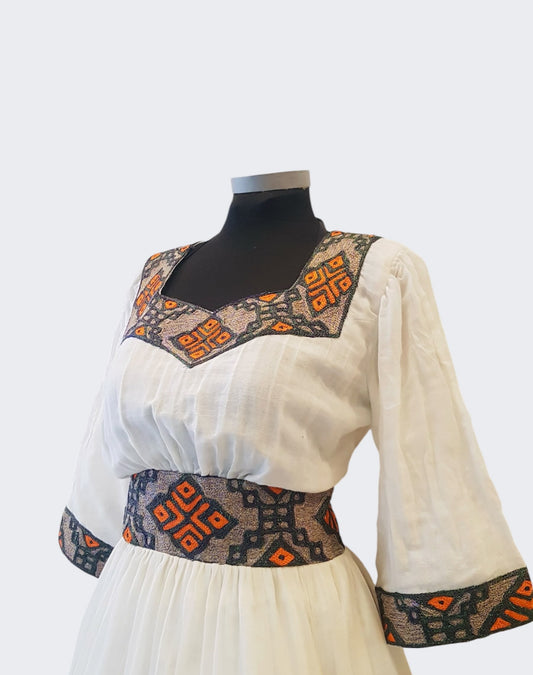 A white long-sleeve ethiopian outfit with geometric patterned trim on the neckline, waist, and cuffs, displayed on a black mannequin torso against a light gray background.u