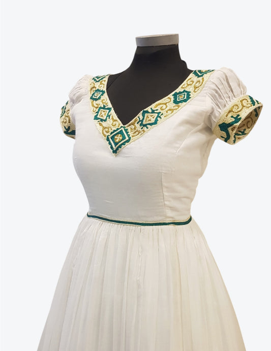 An off-white cotton Ethiopian Outfit displayed on a black mannequin torso, featuring a V-neckline with green and gold embroidered trim, short puff sleeves with matching embroidery, and a green waistband.