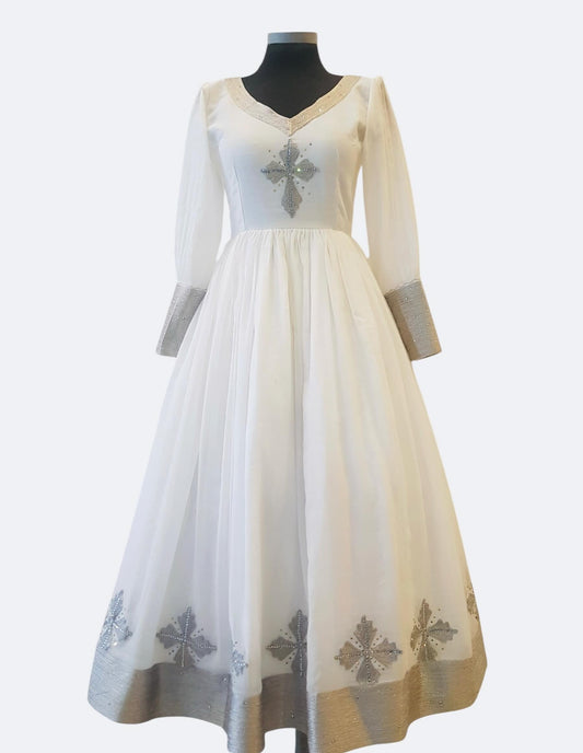A long white or ivory Ethiopian Outfit displayed on a black mannequin against a light gray background. The dress has sheer long sleeves, a V-neckline, a silver embroidered cross on the bodice, and matching silver embroidered motifs near the hem.