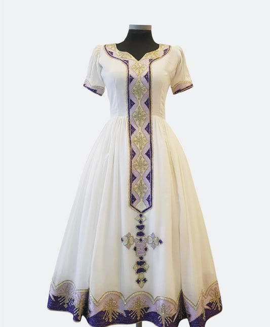 A white A-line Ethiopian Outfit  displayed on a black mannequin against a light gray background. The dress features short sleeves, a V-neckline, and ornate purple and gold embroidery concentrated on the front panel, hem, and a vertical cross motif near the bottom.
