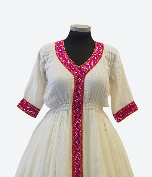 An off-white cotton tunic Ethiopian Outfit  with magenta and purple geometric embroidered trim on the V-neckline, front placket, and sleeve cuffs, displayed on a black mannequin torso against a light gray background.