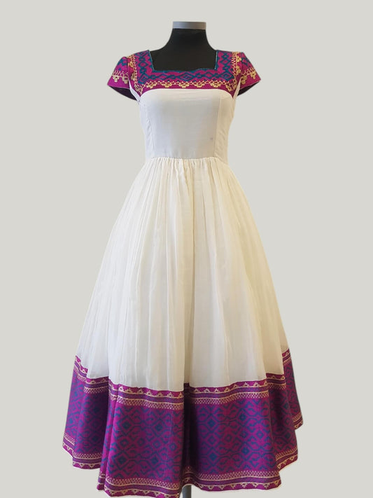 A white A-line Ethiopian outfit  displayed on a black mannequin against a light gray background. The dress features a square neckline with purple and pink patterned embroidery, a flared white skirt, and a matching purple and pink patterned hem.