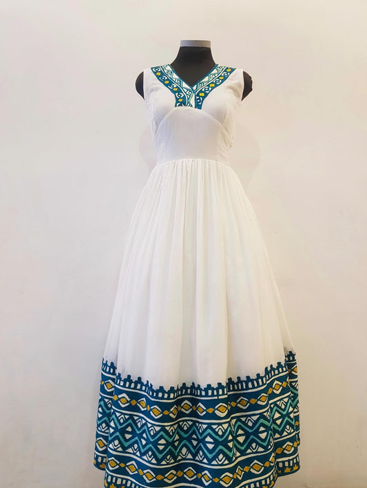 A sleeveless white maxi Ethiopian outfit  displayed on a black mannequin against a plain off-white wall. The dress features a V-neckline with teal and yellow geometric embroidery, and a matching embroidered border along the hem.