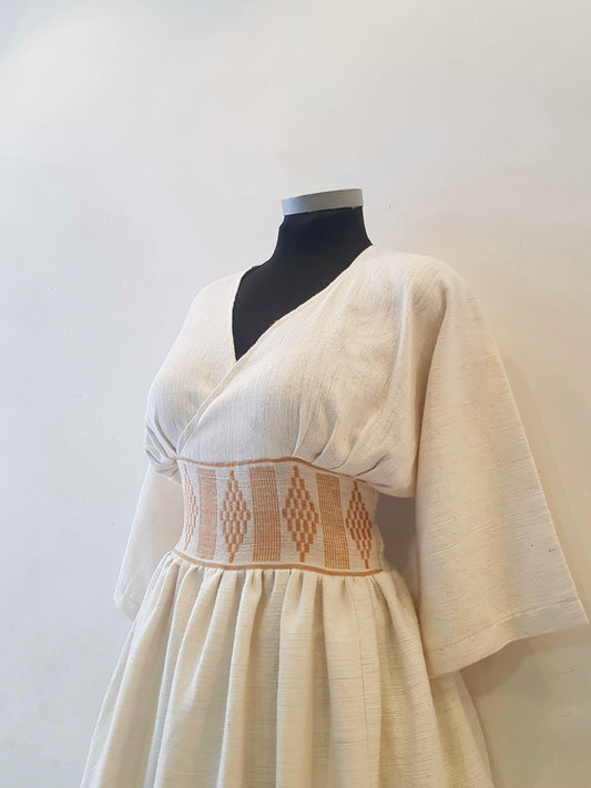 An off-white linen Ethiopian outfit  displayed on a black mannequin torso against a plain light background. The dress features a wrap-style V-neck bodice, wide short sleeves, and a gathered smocked waistband with a tan geometric diamond pattern.