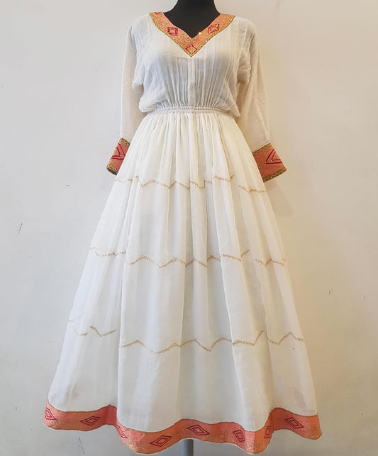 A white traditional long Ethiopian outfit  displayed on a black mannequin against a plain off-white wall. The dress features a V-neckline with orange and gold patterned trim, three-quarter sleeves with matching patterned cuffs, and a full skirt decorated with horizontal gold zigzag lines and an orange patterned hem.