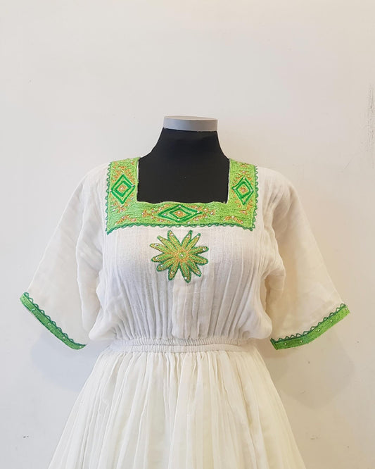 A white cotton Ethiopian outfit  displayed on a black mannequin torso against a plain white wall. The dress features green embroidered geometric patterns around the square neckline and sleeve cuffs, with a green and yellow embroidered sunflower motif on the center chest.