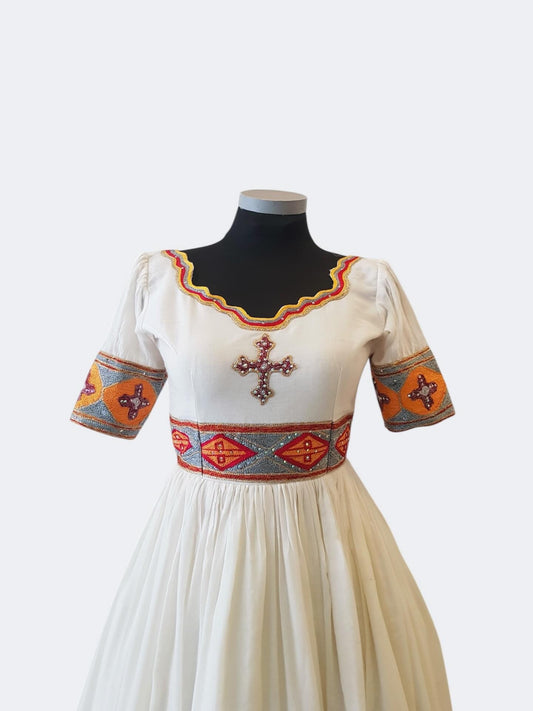 An off-white traditional Ethiopian outfit displayed on a black mannequin torso against a light gray background. The dress features short puff sleeves with orange, red, and blue geometric patterns, a matching patterned waistband, and a central embroidered cross on the chest.