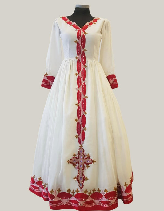 A long white Ethiopian Outfit displayed on a black mannequin against a light gray background. The dress features red embroidered details, including a vertical decorative panel down the front, red cuffs, and a red and gold embroidered cross near the hem.