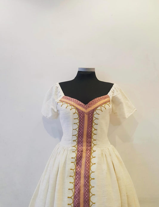 A cream-colored medieval-style Ethiopian Outfit displayed on a black mannequin torso against a plain off-white wall. The dress features short puff sleeves, a fitted bodice, and a gathered skirt, decorated with a central vertical panel and trim in purple, gold, and red.