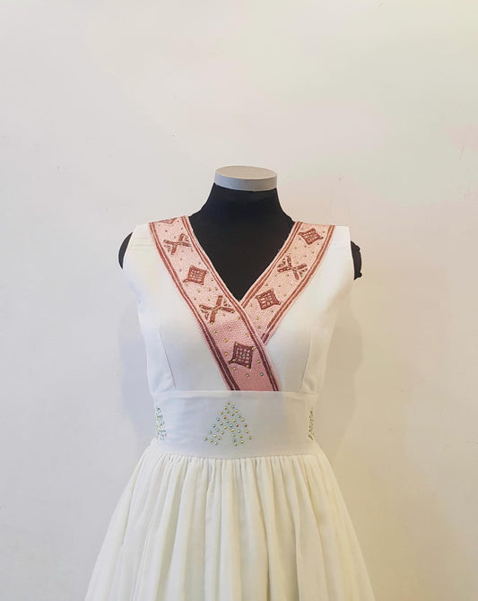 A sleeveless white  Ethiopian Outfit  displayed on a black mannequin torso against a plain off-white wall. The dress has a V-neckline with a wide pink embroidered band across the chest featuring geometric patterns, and small green embroidered details near the waistband.