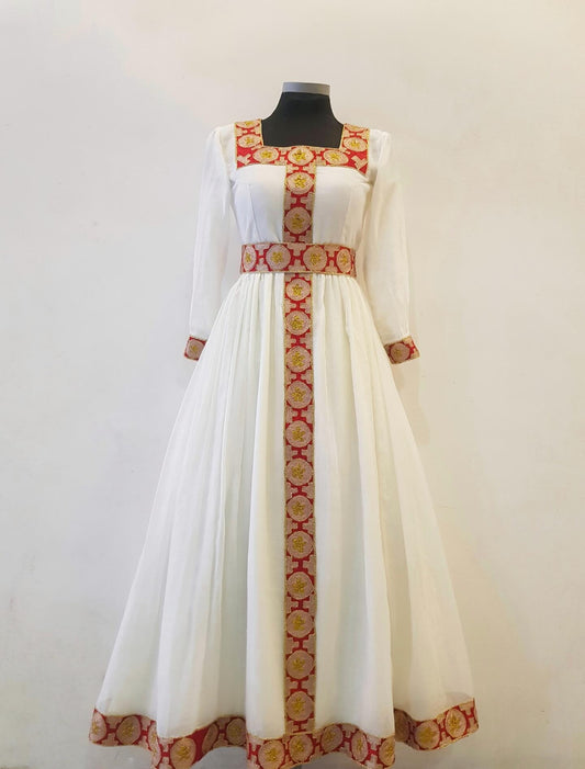 A long white Ethiopian Outfit displayed on a black mannequin against a plain off-white wall. The dress has three-quarter sheer sleeves, a square neckline, and a full skirt, decorated with red and gold patterned trim along the neckline, waist, cuffs, and hem.