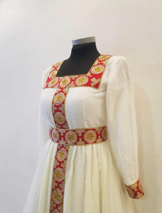 A white long-sleeved Ethiopian Outfit displayed on a black mannequin torso against a plain light background. The dress features ornate red and gold patterned trim along the neckline, waist, and front panel.