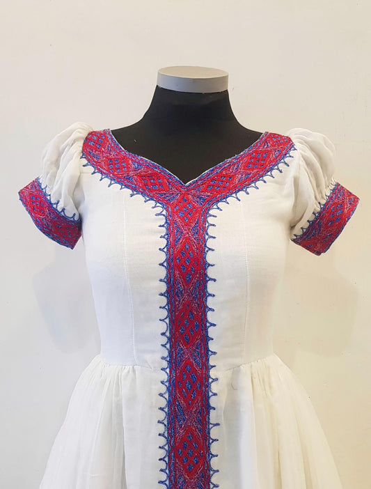 A white traditional-style  Ethiopian Outfit  displayed on a black mannequin torso against a plain light background. The dress features red and blue embroidered trim along the V-neckline, front placket, sleeve cuffs, and puffed short sleeves.