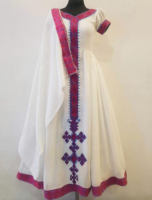 A white traditional  Ethiopian Outfit  displayed on a black mannequin against a plain light beige wall. The dress features vibrant red and blue geometric embroidery along the neckline, sleeve, center front, hem, and a draped matching scarf.
