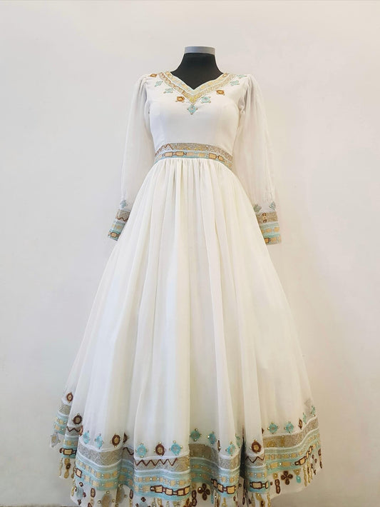 A long white or ivory Ethiopian outfit  displayed on a black mannequin against a plain light wall. The dress has long sheer sleeves, a V-neckline, a fitted waist with a decorative embroidered band, and a full flowing skirt with matching embroidered trim near the hem.