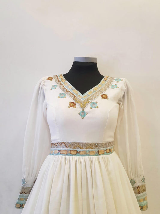 A white long-sleeved Ethiopian outfit  displayed on a black mannequin torso against a plain light background. The dress features a V-neckline with gold and turquoise embroidery, a matching embroidered waistband, and detailed gold and turquoise patterns on the cuffs.