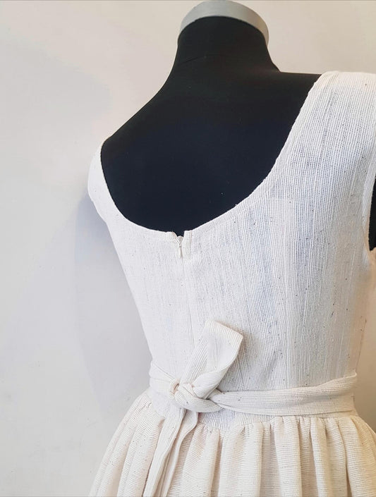 A close-up of a white textured sleeveless Ethiopian outfit  displayed on a black mannequin torso, showing the back view with a scoop neckline, tie detail at the waist, and gathered skirt.