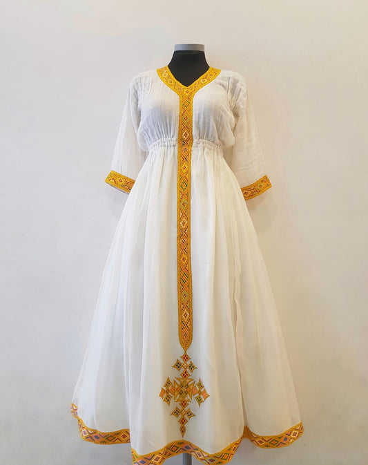 A white A-line Ethiopian outfit  displayed on a black mannequin against a plain light background. The dress features gold embroidered details, including a vertical panel down the front, sleeve cuffs, and a geometric border along the hem.