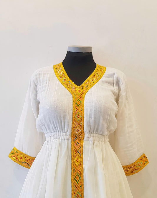 A white cotton tunic Ethiopian outfit  with yellow and orange geometric embroidered trim on the V-neckline, front placket, cuffs, and waist, displayed on a black mannequin torso against a plain off-white wall.