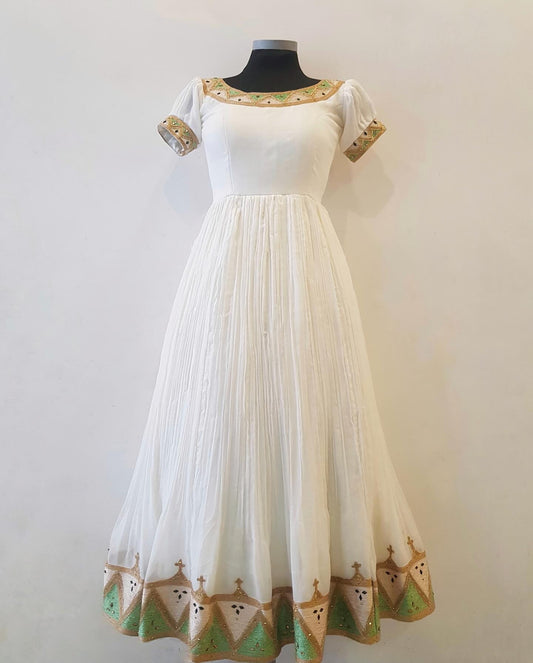 A white A-line maxi Ethiopian outfit  with short puff sleeves, a square neckline, and decorative gold and green embroidered borders, displayed on a black mannequin torso against a plain off-white wall.