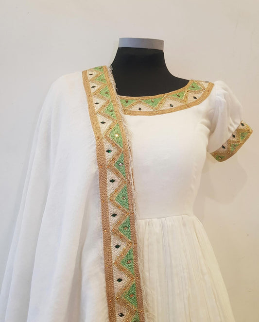 A white traditional Ethiopian outfit  displayed on a black mannequin torso, featuring a sheer white dupatta draped over the shoulder with a wide decorative border in beige, green, and black geometric embroidery.