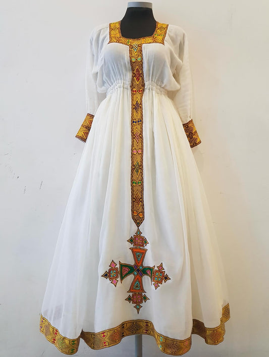 A white long-sleeved Ethiopian outfit  displayed on a black mannequin torso against a plain white wall. The dress features ornate multicolor embroidery in gold, red, green, and blue, concentrated around the neckline, cuffs, and a large cross motif on the skirt.