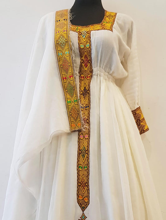 A white traditional Ethiopian outfit displayed on a black mannequin against a plain light background. The garment features prominent gold, orange, and green geometric embroidered borders along the neckline, front opening, and sleeve cuffs.
