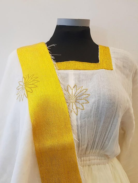 A white embroidered Ethiopian outfit  with a wide mustard yellow sash draped over the shoulder, displayed on a black mannequin torso against a plain off-white background.