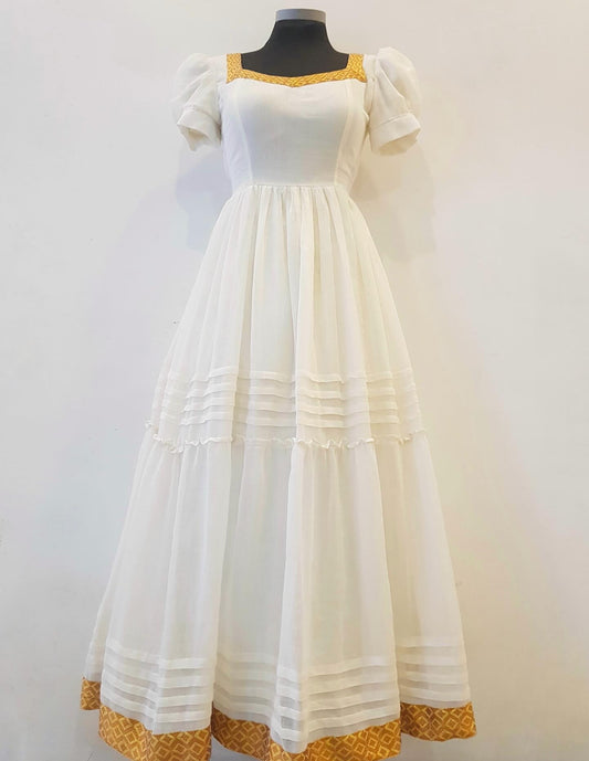 A white tiered maxi Ethiopian outfit  with puff sleeves and gold patterned trim at the neckline, armholes, and hem, displayed on a black mannequin torso against a plain white wall.