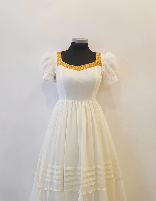 A white A-line Ethiopian outfit  with puff sleeves and a gold embroidered neckline, displayed on a black mannequin torso against a plain off-white wall.