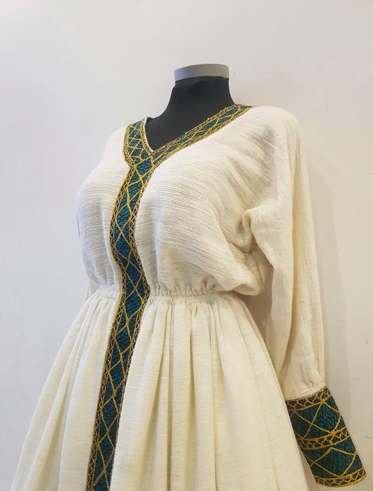 A cream-colored medieval-style Ethiopian outfit  displayed on a black mannequin torso. The dress features a V-neckline and long sleeves, both adorned with a teal and gold geometric patterned trim that runs down the front and onto the cuffs.