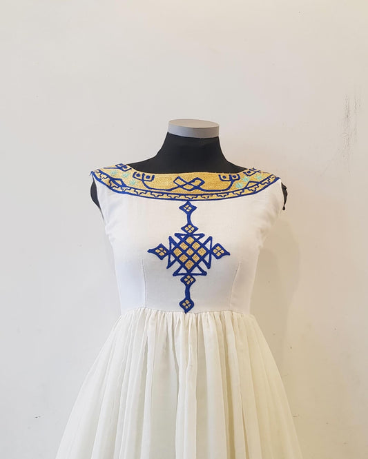 An off-white sleeveless Ethiopian Outfit displayed on a black mannequin torso against a plain light wall. The dress features a wide yellow and blue embroidered yoke and a large blue embroidered cross motif on the front bodice, with a gathered off-white skirt.