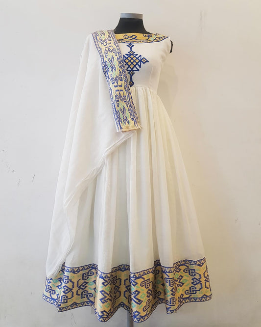 A white traditional Ethiopian outfit  displayed on a black mannequin against a plain off-white wall. The dress features a white body, a draped white dupatta with blue and yellow geometric borders, and a wide yellow border along the hem with blue geometric patterns.