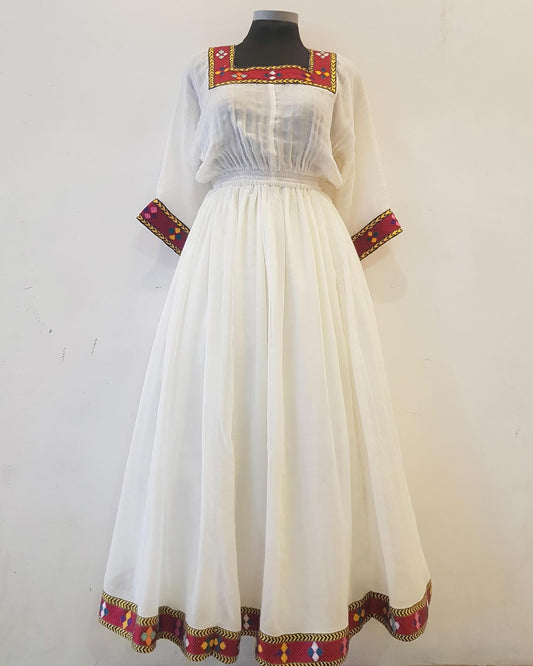 A long white Ethiopian outfit displayed on a black mannequin against a plain off-white wall. The dress has sheer long sleeves, a gathered bodice, and ornate embroidered borders in red, gold, and green along the neckline, cuffs, and hem.