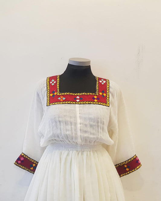 A white traditional-style Ethiopian outfit displayed on a black mannequin torso against a plain off-white wall. The dress features a square neckline with a red embroidered border and multicolored geometric motifs, long sheer sleeves with matching embroidered cuffs, and a gathered white skirt.