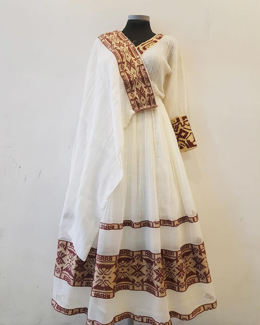 A white traditional Ethiopian outfit displayed on a black mannequin against a plain off-white wall. The outfit features brown and gold geometric patterns along the neckline, sleeve cuffs, and hem, with a matching sheer white dupatta draped over the shoulders.