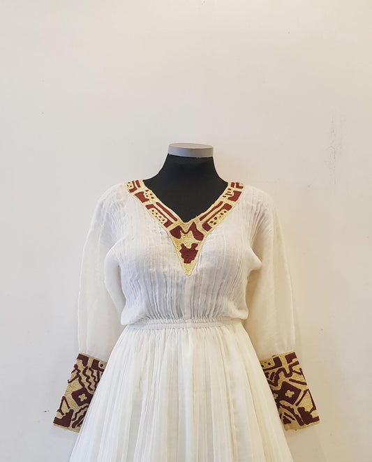 A white long-sleeve tunic Ethiopian outfit with brown and gold geometric embroidery on the V-neckline and cuffs, displayed on a black mannequin torso against a plain off-white wall.