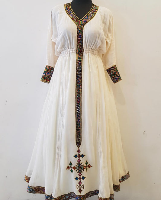 An off-white maxi Ethiopian outfit displayed on a black mannequin against a plain white wall. The dress features colorful geometric embroidered details along the V-neckline, cuffs, center placket, and a matching cross motif near the hem.