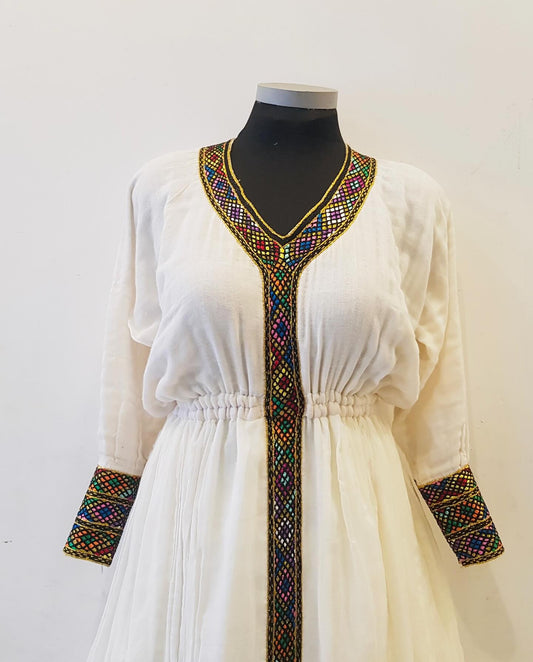 A white long-sleeve Ethiopian outfit displayed on a black against a plain off-white background. The dress features a V-neckline and gathered waist with vibrant, multicolored geometric embroidery running down the center front and along the cuffs.