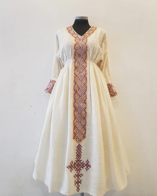 An off-white linen Ethiopian outfit with red and purple geometric embroidery, displayed on a black mannequin against a plain white wall.