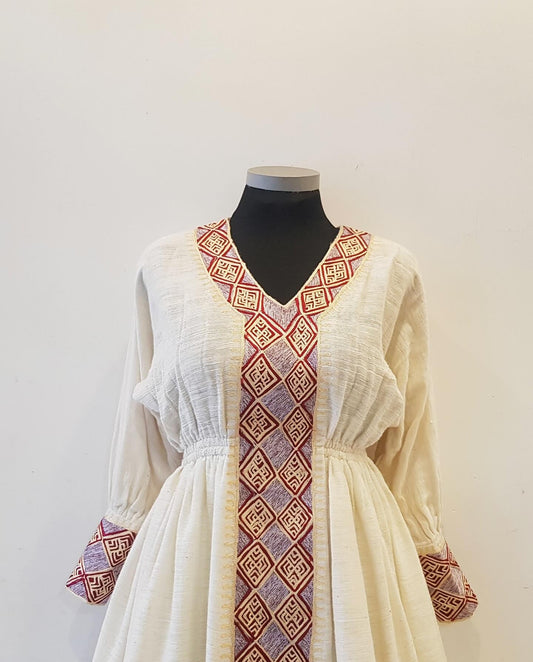 An off-white linen long-sleeve Ethiopian outfit with red and purple geometric embroidered trim, displayed on a black mannequin torso against a plain white wall.