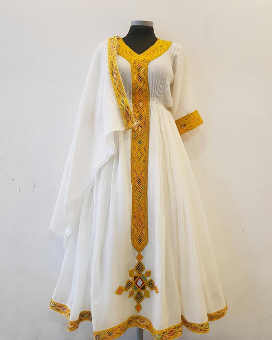 A white traditional Ethiopian outfit displayed on a black mannequin against a plain off-white wall. The dress features prominent yellow and orange geometric embroidery along the neckline, sleeves, hem, and a vertical chest panel.