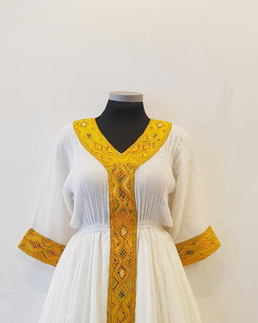 A white traditional Ethiopian outfit displayed on a black mannequin torso against a plain off-white wall. The dress features a deep V-neckline and wide sleeves, both adorned with prominent yellow embroidered lace trim and a matching vertical panel down the center.