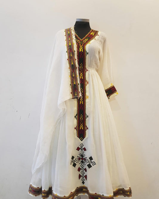 A white traditional long Ethiopian outfit displayed on a black mannequin against a plain off-white wall. The dress features prominent dark red, yellow, and green geometric embroidery along the V-neckline, sleeve cuffs, and a matching border at the hem, with a central embroidered motif near the lower skirt.